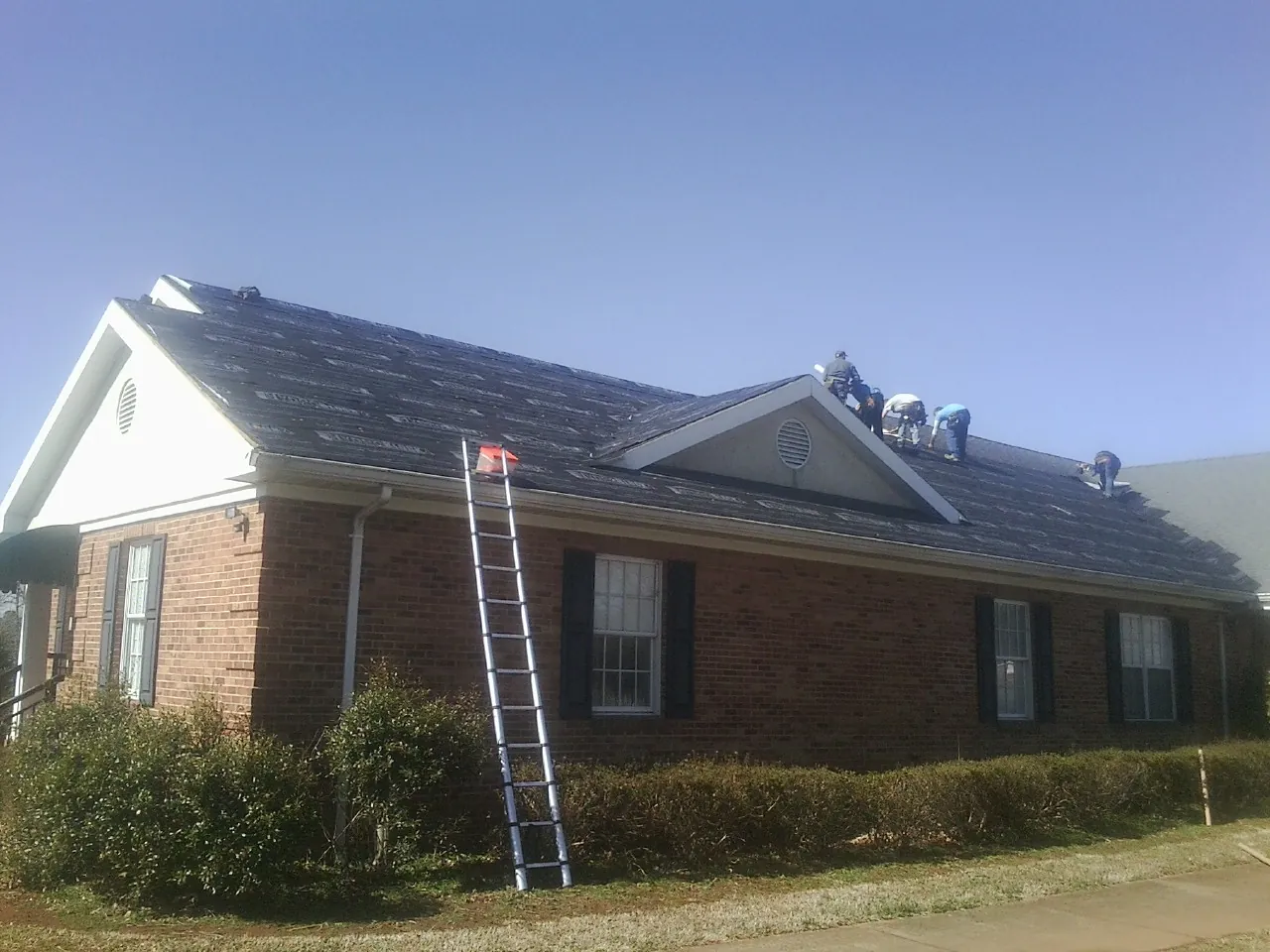 Crew tearing shingles off a brick ranch roof with a ladder set — Emergency Roof Repair work by our crew in Pleasant Hill