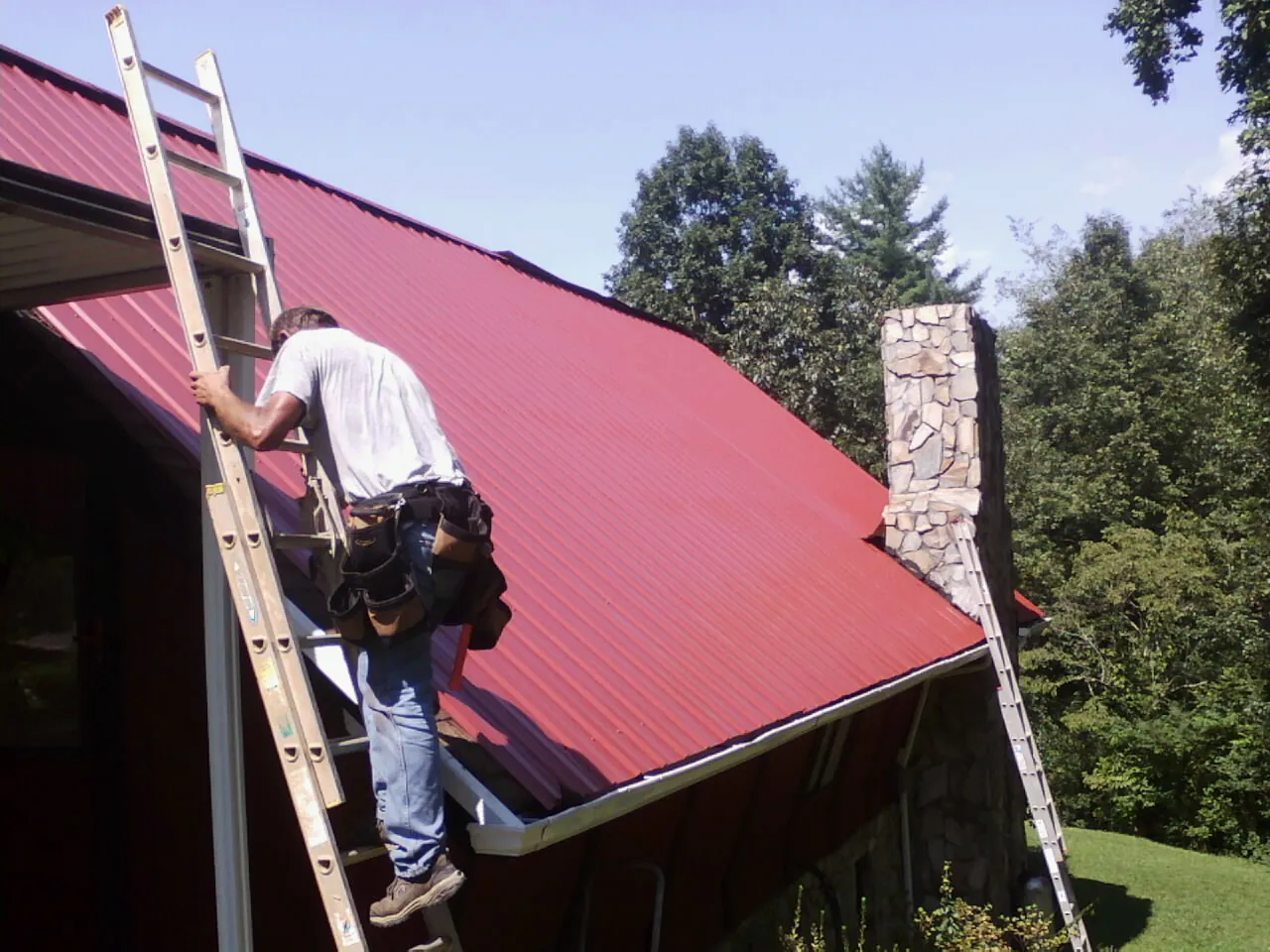 Roofer on a ladder beside a red metal roof and stone chimney — Soffit & Fascia Repair by Roofing Brothers in Pleasant Hill